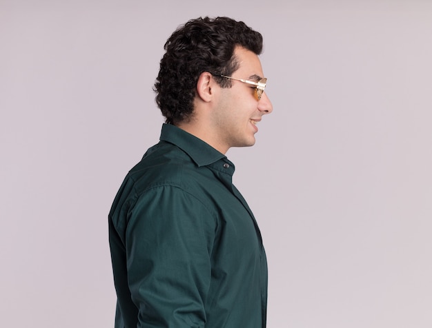 Young man in green shirt wearing glasses standing sideways with smile on face over white wall