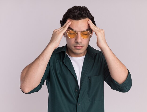 Молодой человек в зеленой рубашке в очках трогает голову усталым и перегруженным работой с головной болью, стоящей над белой стеной