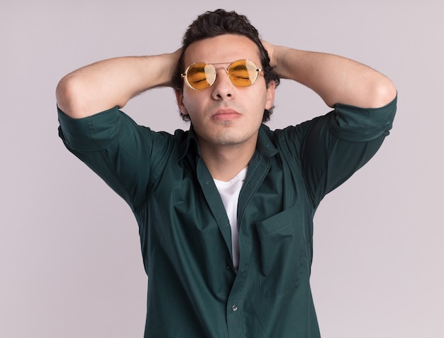 Young man in green shirt wearing glasses with closed eyes annoyed with hands behind his head standing over white wall