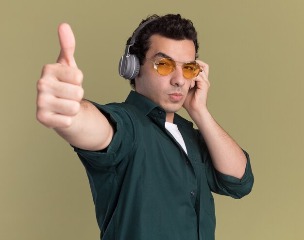 緑の壁の上に立って親指を示す深刻な顔で正面を見てヘッドフォンで眼鏡をかけている緑のシャツの若い男