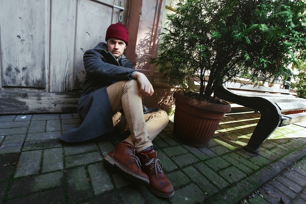 Young man in grey coat and beige pants sits on the pavement 