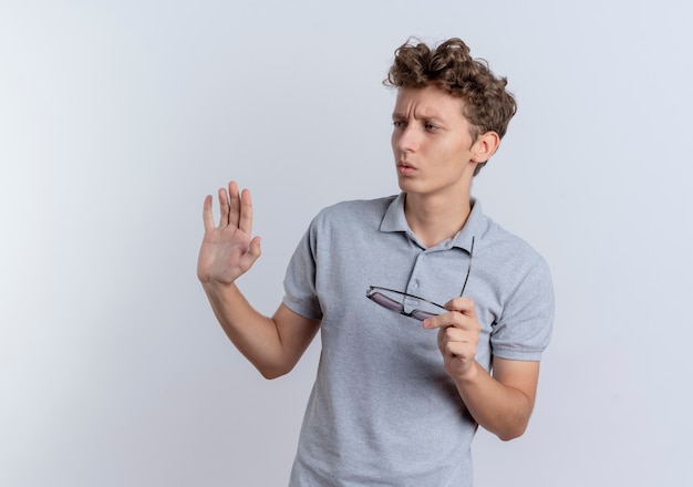 Young man in grey polo shirt holding glasses looking aside with confuse expression standing over white wall