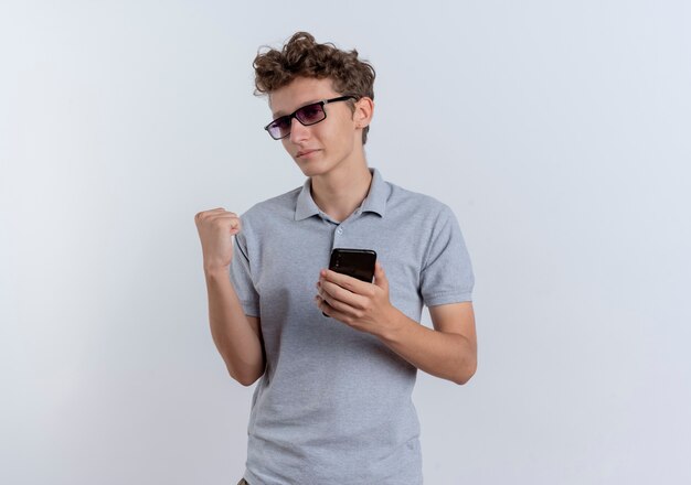 白い壁の上に立って幸せで興奮して拳を握りしめるスマートフォンを保持している灰色のポロシャツの若い男