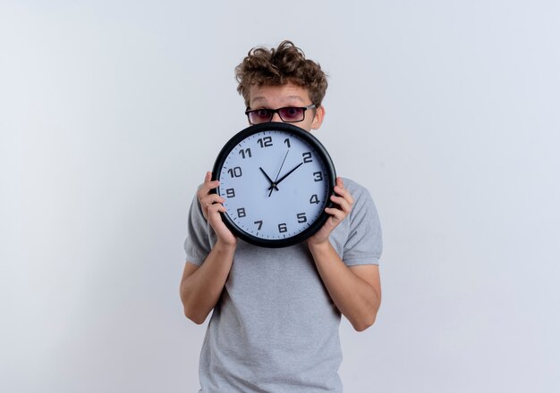 Young man in grey polo shirt holding wall clock hiding face peeking over surprised standing over white wall