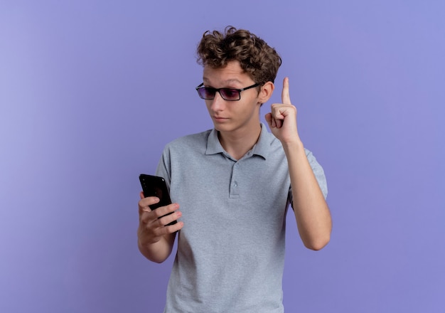 Young man in grey polo shirt looking at his smartphone screen being surprised and happy showing index finger having new idea standing over blue wall