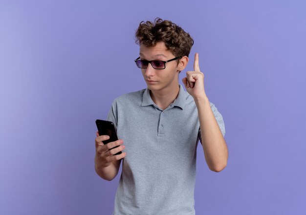Молодой человек в серой рубашке поло смотрит на экран своего смартфона, удивленный и счастливый, показывая указательный палец, имеющий новую идею, стоящий над синей стеной