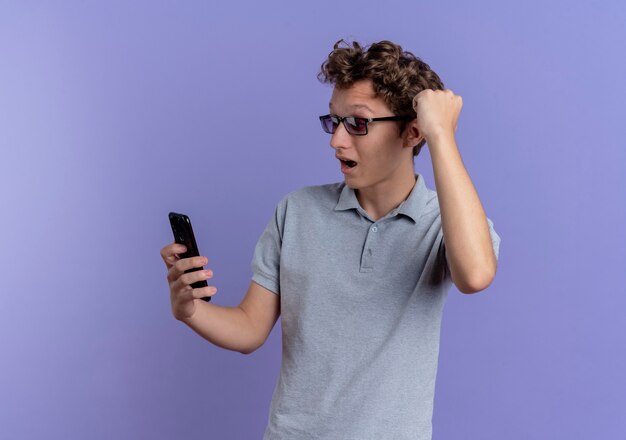 Young man in grey polo shirt looking at his smartphone screen clenching fist happy and excited standing over blue wall