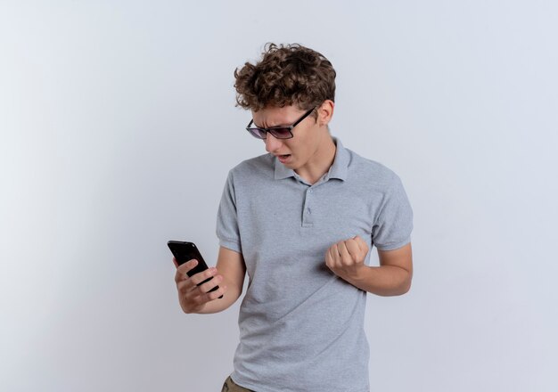 Молодой человек в серой рубашке поло смотрит на экран своего смартфона, сжимая кулак, счастлив и взволнован, стоя над белой стеной