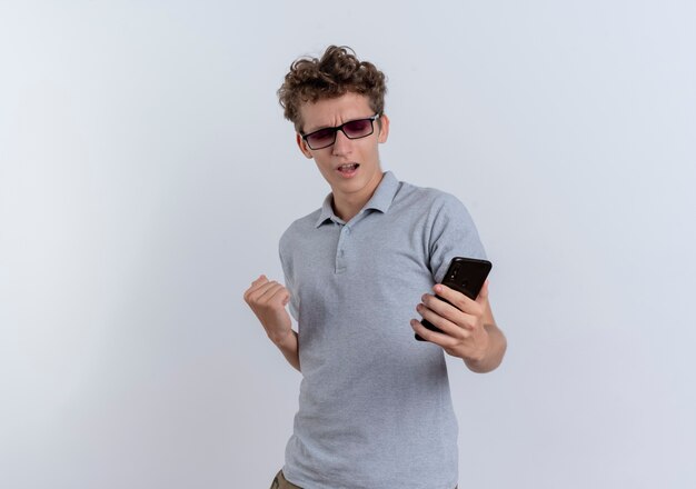 Young man in grey polo shirt looking at his smartphone screen clenching fist rejoicing his success standing over white wall