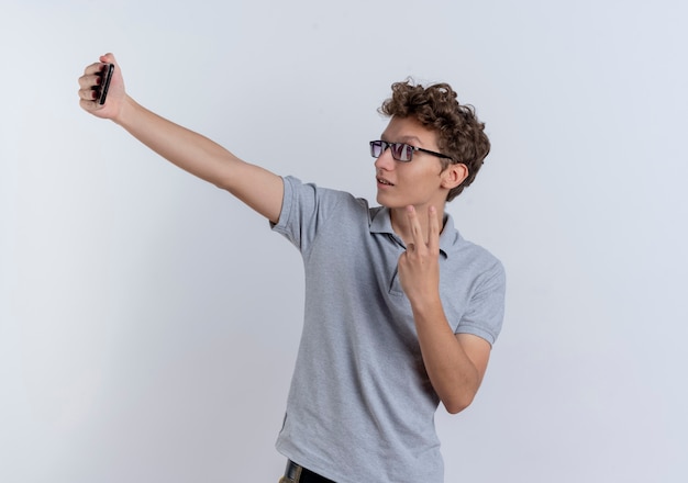 Young man in grey polo shirt looking at his smartphone screen doing selfie showing v-sign standing over white wall