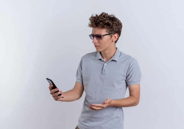 Молодой человек в серой рубашке поло, глядя на экран своего смартфона, выглядит смущенным, стоя над белой стеной