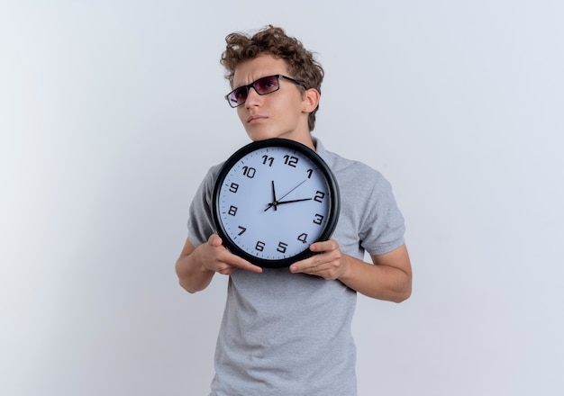 Young man in grey polo shirt showing wall clock looking aside confused standing over white wall