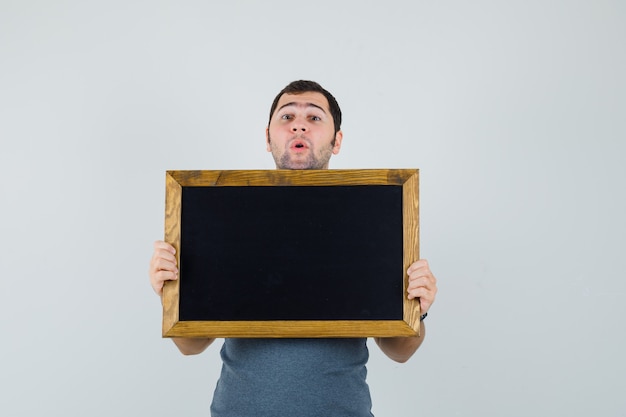 Young man in grey t-shirt holding blackboard and looking amazed  