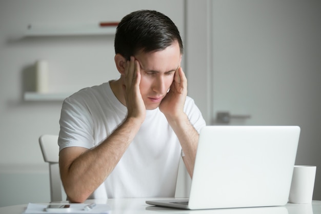 Young man, hands at his temples, at the white desk, laptop near