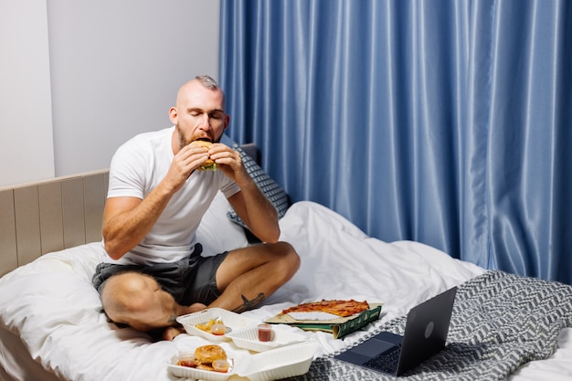 Young man having fast food at home in bedroom on bed
