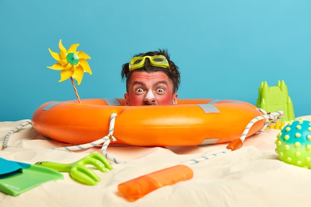 Голова молодого человека с солнцезащитным кремом на лице в окружении пляжных аксессуаров