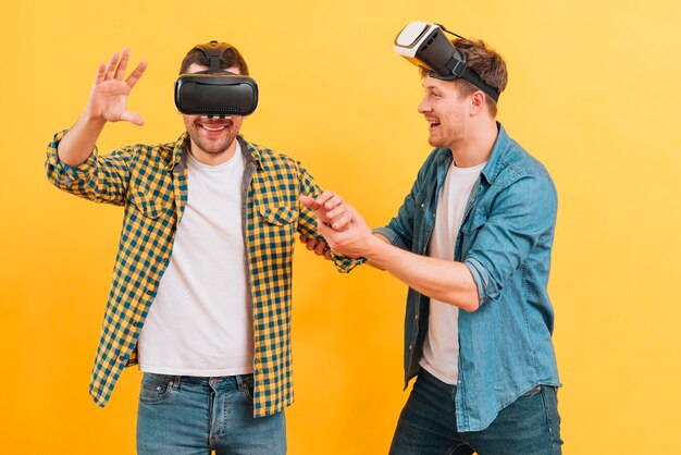 Free photo young man helping his friend wearing glasses of virtual reality against yellow background