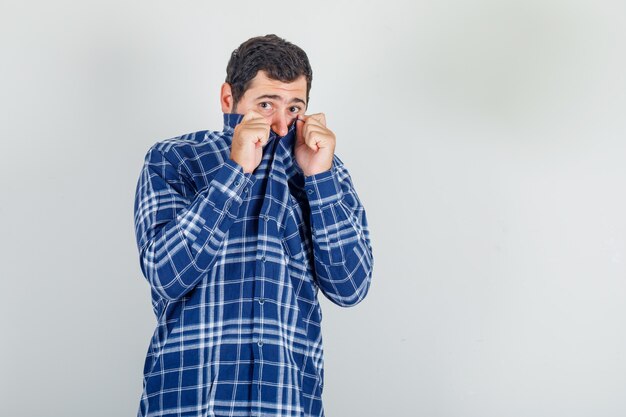 young man hiding face behind collar in checked shirt and looking scared.