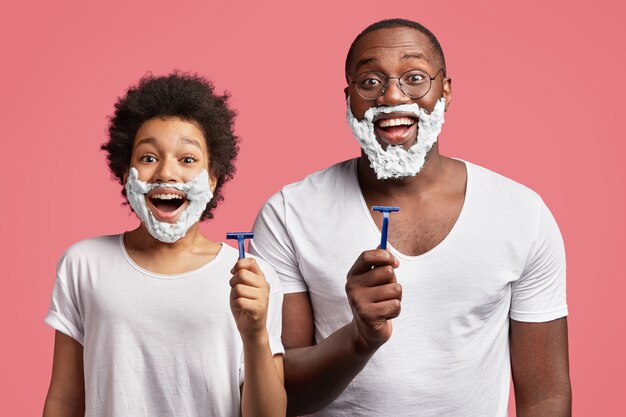 Free photo young man and his son having shaving foam on face and holding razors