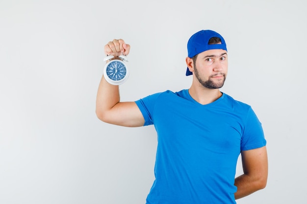 Молодой человек держит будильник в синей футболке и кепке и смотрит задумчиво