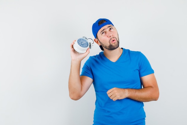 Young man holding alarm clock to hear noise in blue t-shirt and cap front view.