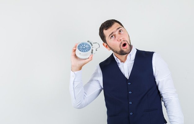 Young man holding alarm clock near ear in shirt, vest and looking curious