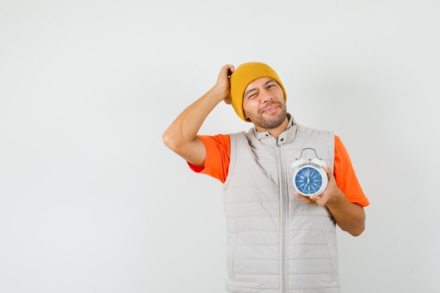 Young man holding alarm clock, scratching head in t-shirt, jacket, hat and looking forgetful. front view.