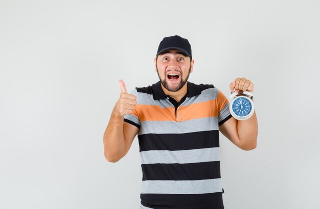 Young man holding alarm clock, showing thumb up in t-shirt, cap and looking happy. front view.