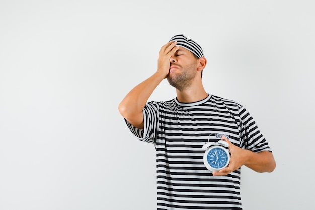 Young man holding alarm clock in striped t-shirt hat and looking forgetful  