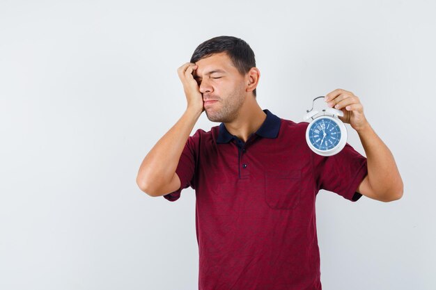 Young man holding alarm clock in t-shirt and looking forgetful. front view.