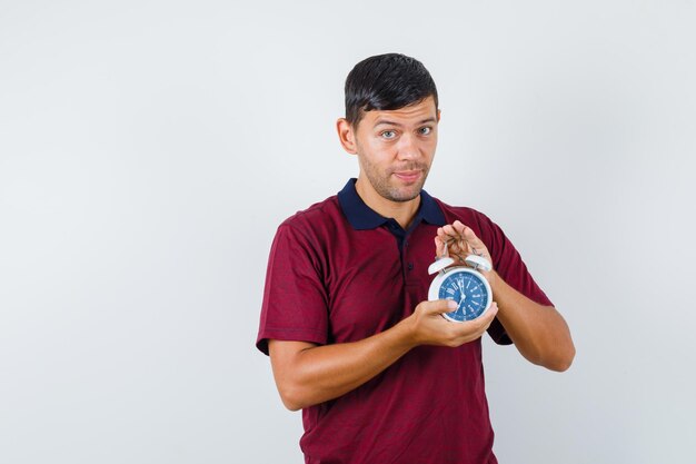 Young man holding alarm clock in t-shirt and looking positive. front view.