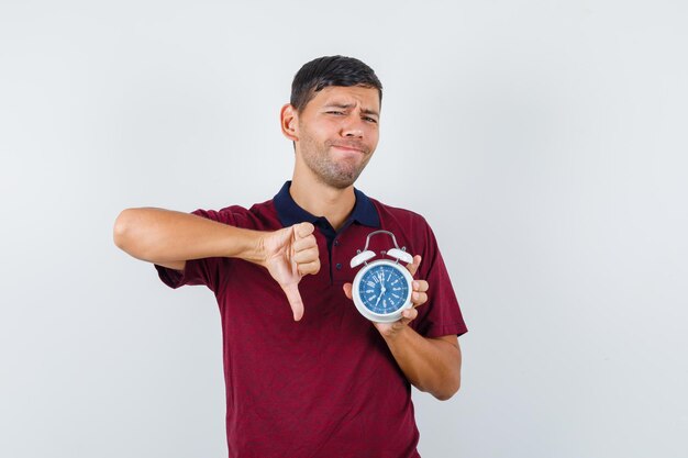 Young man holding alarm clock with thumb down in t-shirt and looking displeased , front view.