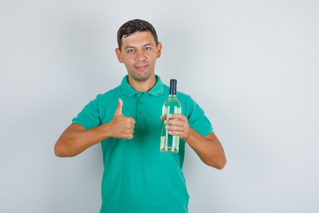 Young man holding alcohol bottle and showing thumb up in green t-shirt and looking pleased, front view.