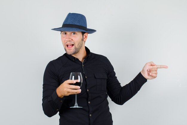 Young man holding alcohol glass while pointing to side in black shirt