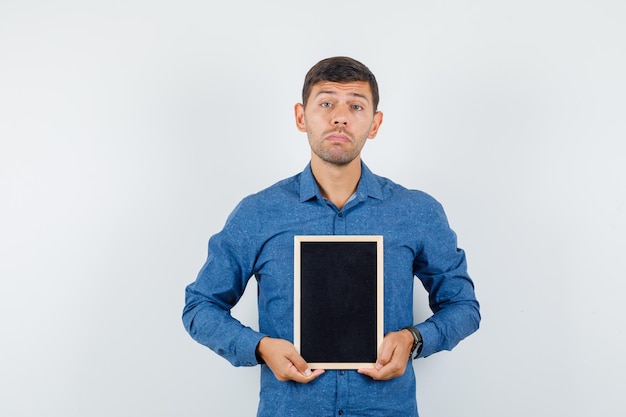 Young man holding blackboard in blue shirt and looking helpless. front view.