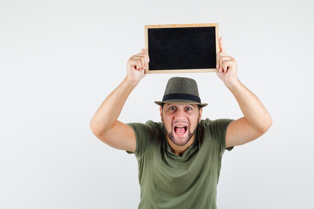 Young man holding blackboard over head in green t-shirt and hat and looking lively