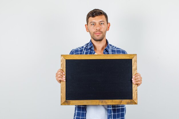 Young man holding blackboard in shirt and looking confident. front view.