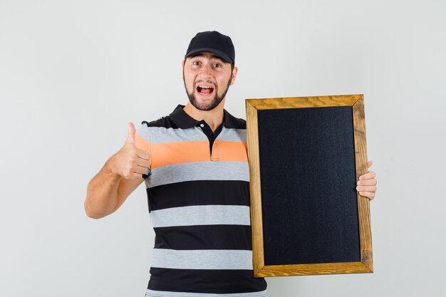 Young man holding blackboard, showing thumb up in t-shirt, cap and looking happy. front view.