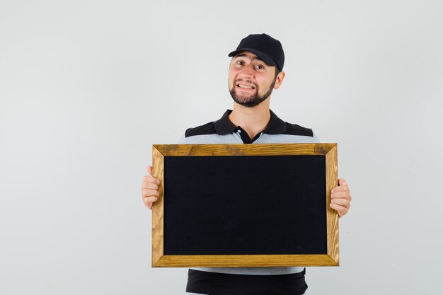 Young man holding blackboard in t-shirt, cap and looking pleased , front view.