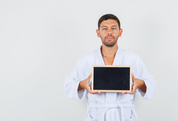Young man holding blackboard in white bathrobe and looking serious. front view.