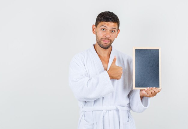 Young man holding blackboard with thumb up in white bathrobe front view.