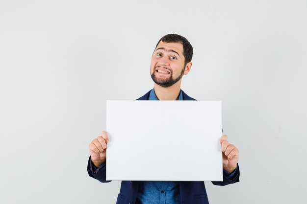 Young man holding blank canvas in shirt, jacket and looking merry. front view.