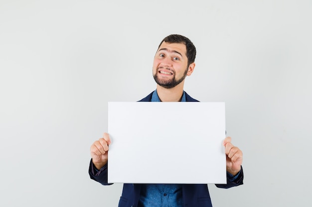 Young man holding blank canvas in shirt, jacket and looking merry. front view.