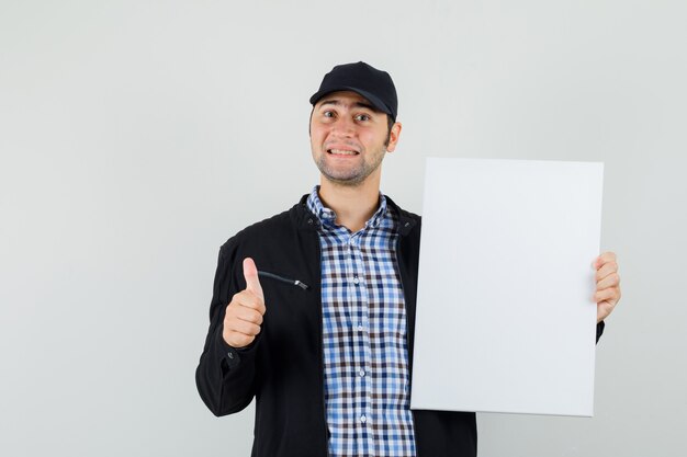 Young man holding blank canvas, showing thumb up in shirt, jacket, cap and looking optimistic. 