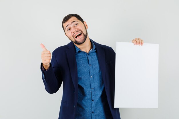 Young man holding blank canvas, showing thumb up in shirt, jacket and looking merry. front view.