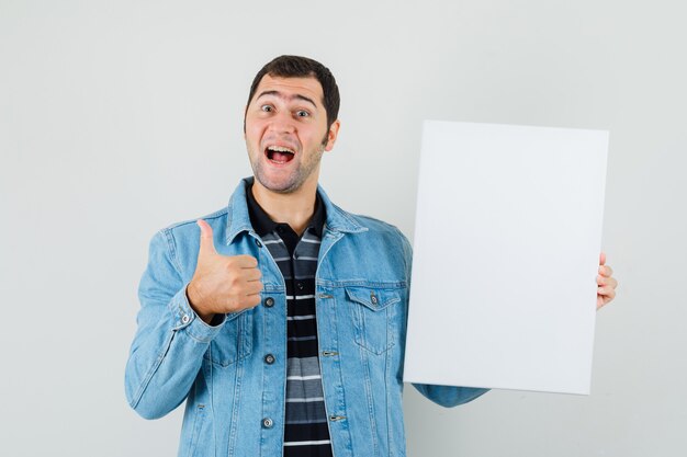 Young man holding blank canvas, showing thumb up in t-shirt, jacket and looking cheerful