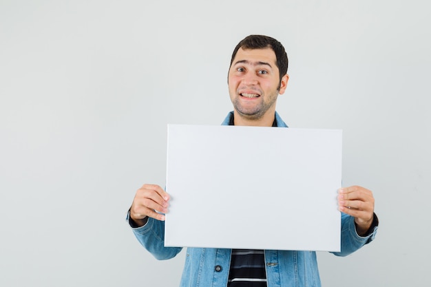 Young man holding blank canvas in t-shirt, jacket and looking merry