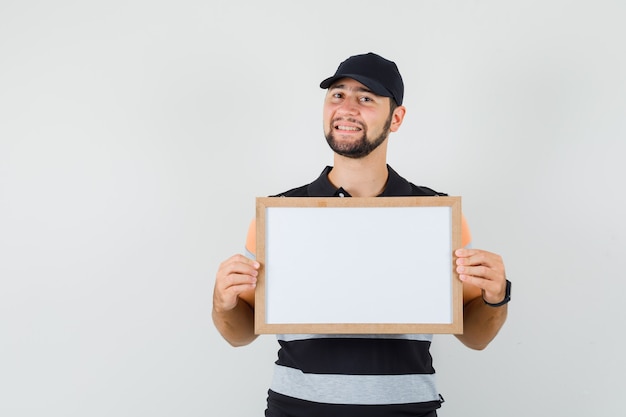 Young man holding blank frame in t-shirt, cap and looking cheerful , front view.