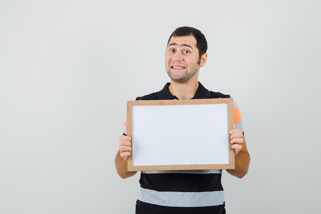 Young man holding blank frame in t-shirt and looking attentive.  space for text