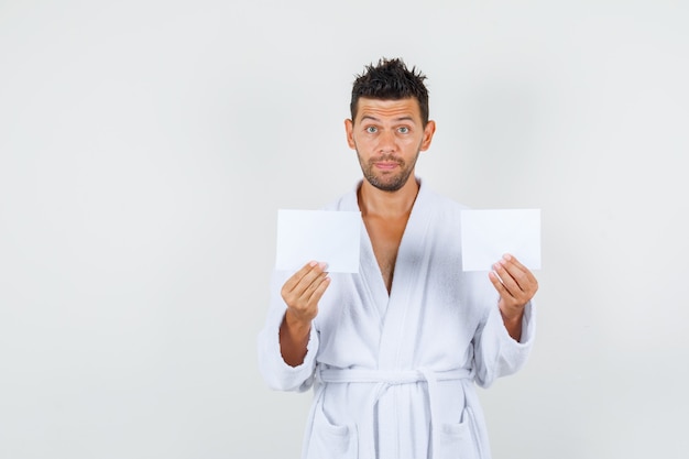 Young man holding blank paper sheets in white bathrobe front view.